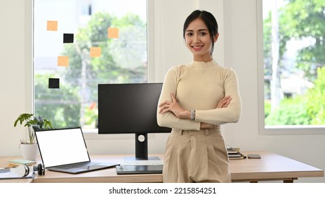 Attractive And Beautiful Millennial Asian Businesswoman Or Female CEO In Casual Clothes Arms Crosses, Smiling, Looking At Camera  While Standing In Her Modern Minimal Office.