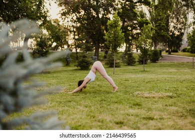 Attractive, Beautiful Woman Does Yoga And Stands In Downward-facing Dog Pose On Green Grass Of Lawn At Sunset, Concept Of Inner Harmony.