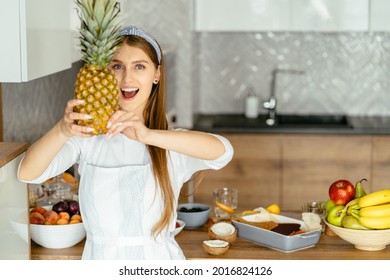 Attractive beautiful woman wearing white shirt, apron holding pineappe and hiding half of her face, looking at camera with happy smile while standing in modern kitchen at home. Healthy food concept - Powered by Shutterstock