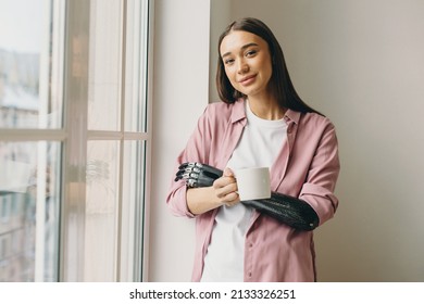 Attractive Beautiful Youthful Brunette Girl With Bionic Black Prosthetic Hand Holding Mug Of Coffee Standing Next To Huge White Window, Admiring Beauty Of Morning, Looking At Camera With Cute Smile