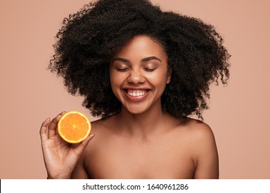 Attractive black female with clean skin laughing with closed eyes and showing half of fresh orange while advertising benefits of vitamin C, in skincare industry against brown background