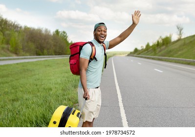 Attractive Black Guy With Luggage Waving Down A Car, Hitchhiking On Roadside, Traveling By Autostop, Outdoors. Handsome African American Man Hitching His Ride, Going On Road Trip, Copy Space