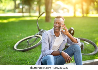 Attractive Black Guy Sitting On Ground Near His Bike And Speaking On Cellphone At Park