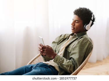 Attractive Black Teen In Headset Choosing Music Playlist On Cellphone At Home. Funky African American Guy Downloading His Favorite Songs On Mobile Device, Listening To Audiobook Or Radio