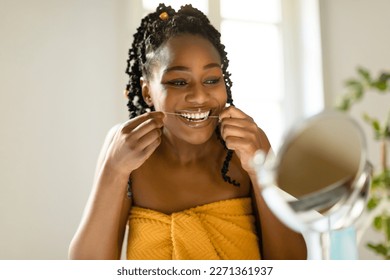 Attractive black woman looking at mirror and using dental floss, cleaning her white teeth, sitting at vanity table in bedroom interior, copy space. Dental care concept