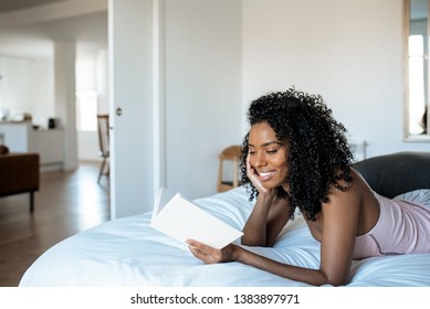Attractive black woman lying down on bed reading a book at home