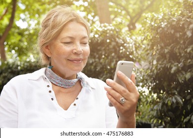 Attractive Blond Elderly Woman Dressed Casually Sitting On Bench In Park Using Smart Phone, Reading Messages From Her Granddaughter Who Graduated Successfully From University With Proud Expression