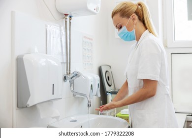 Attractive blond female lab assistant in sterile uniform and with protective mask on face standing in rest room and washing her hands during covid outbreak.