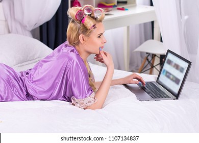 Attractive Blond Female Watching Youtube In Bed. Goodlooking Girl Wearing Lilac Robe Watching Videos On Laptop On White Blanket In Her Bedroom.