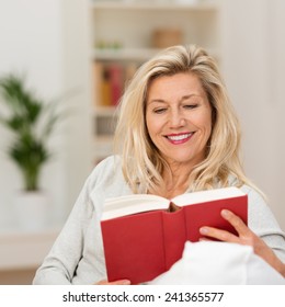 Attraktive blond mittelalterliche Frau, die ein Buch auf dem Sofa in ihrem Wohnzimmer lächelnd lächelt, wie sie liest