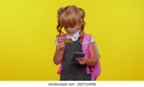 Attractive Blond Teenage Girl Kid In School Uniform Using Credit Bank Card And Smartphone While Transferring Money, Purchases Online Shopping Isolated On Yellow Background. Education, Back To School