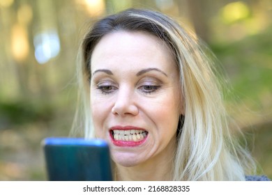 Attractive Blond Woman Examining Her Teeth In A Hand Mirror Outdoors In A Close Up Head Shot