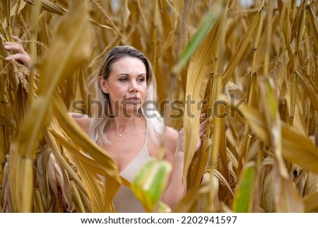 Attractive blond woman wearing a gold dress is standing in the middle of a corn field looking aside depp in thought