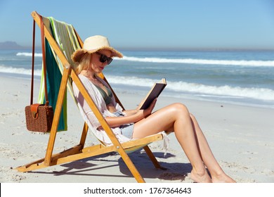 Attractive blonde Caucasian woman enjoying time at the beach on a sunny day, sitting on deck chair, reading a book, with blue sky and sea in the background. Summer tropical beach vacation.