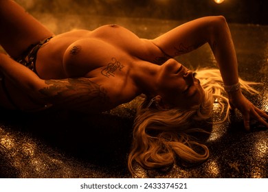 Attractive blonde female posing topless in strapy leather bdsm outfit with whip in front of burlesque light wall with smoke in the air