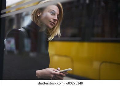 Attractive blonde female tourist waiting for public transport on bus stop using application for navigating in city, charming hipster girl browsing information for traffic in town via smartphone - Powered by Shutterstock