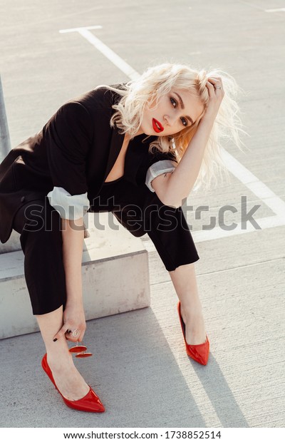 Attractive blonde in a formal\
suit and red shoes in a parking lot near a shopping center at\
sunset\
