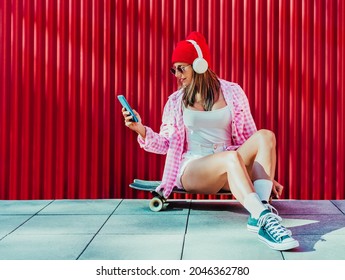 an attractive blonde girl, looking at cell phone messages and listening to music sitting on her skateboard. the background is bright red. - Powered by Shutterstock