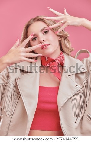 An attractive blonde girl in a white leather jacket and pink top poses against a pink studio background, showing off her stunning pink manicure. Feminine style. Beauty and fashion.