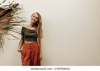 Attractive Blonde Long-haired Happy Woman In Cropped Khaki Top And Red Loose Pants Smiles And Poses Near White Wall With Palm Leaf.