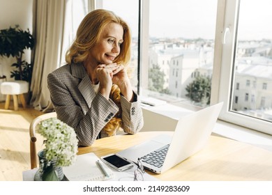 Attractive Blonde Mature Female CEO In Stylish Blazer Sitting At Desk Near Window Placing Chin On Hands Talking To Director General Via Laptop, Phone With Copy Space For Your Text On Table Next To Her