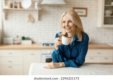 Attractive blonde mature lady drinking fresh coffee or tea at kitchen alone, looking aside and smiling, free space. Smiling woman holding cup, standing in the cozy kitchen - Powered by Shutterstock