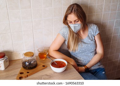 Attractive Blonde Is Sitting In The Kitchen Near The Table With Food. She Is Isolated At Home During Quarantine Due To The Coronavirus Epidemic, Wearing A Protective Mask.Looking To Plate With Soup