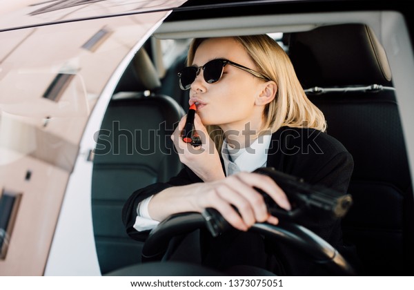 attractive blonde woman applying lipstick while holding\
gun in car 
