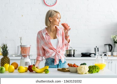 attractive blonde woman drinking tasty smoothie near ingredients in kitchen  - Powered by Shutterstock