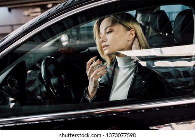 attractive blonde woman holding bottle while spraying perfume in car 
