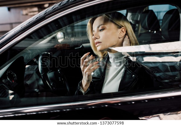 attractive blonde woman holding bottle while spraying\
perfume in car 