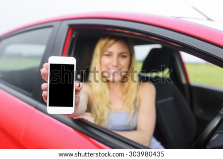 Attractive blonde woman showing smartphone with empty screen out the window of a car. Focus on model.