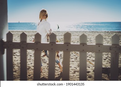 Attractive Blonde Woman Wearing Sunglasses And Casual Outfit Passing By With The Cute Pet Dog Behind The Fence On The Beach On A Sunny Day. Beautiful Female Walking With A Cute Puppy On A Sandy Beach.