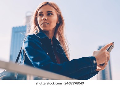 Attractive blonde young woman in denim jacket looking away while installing app on smartphone standing on street.Charming female tourist updating profile in social networks on phone via internet