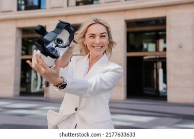 Attractive Blonde Young Woman In White Suite Offering To Try A Vr Glasses, Excited After Virtual Reality Experience. Smiley Student Girl In White Suit Outdoor At Break. Technologies And Entertainment.