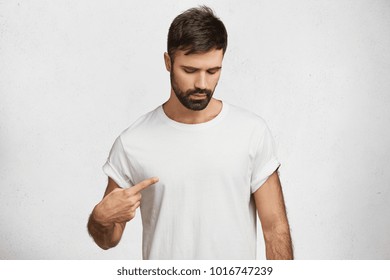 Attractive brunet male with beard, looks seriously down, indicates at blank copy space of t shirt for your logo or advertising text, isolated over white background. People, clothing, design concept