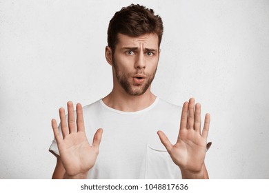 Attractive brunet male shows refusal gesture, doesn`t want to participate in meeting, says: It`s not for me, leave me in piece, has angry expression, poses against white concrete studio background - Shutterstock ID 1048817636