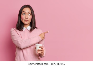 Attractive brunette Asian woman holds disposable cup of coffee points aside and shows blank space advertises place for promotion dressed in casual clothes suggests visit cafe and grab some drink