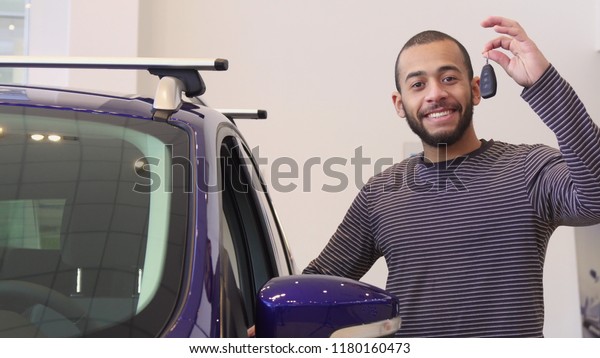Attractive brunette bearded man standing near the\
purple crossover at the dealership. Young african american guy\
examining interior of the car. Handsome male customer demonstrating\
car key at the