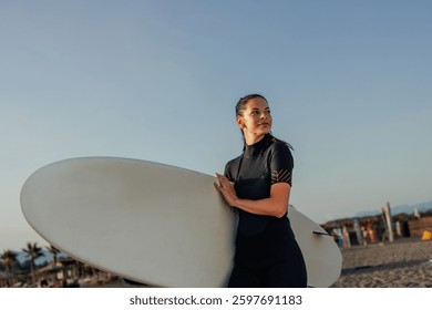 Attractive brunette female surfer wearing a black wet suit is carrying a white surfboard on a sandy beach illuminated by the sun.