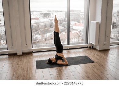 Attractive brunette female in tight top and yoga pants balancing in Shoulder Stand Pose on black mat in training room of sports studio. Active slim woman improving body strength using Sarvangasana. - Powered by Shutterstock