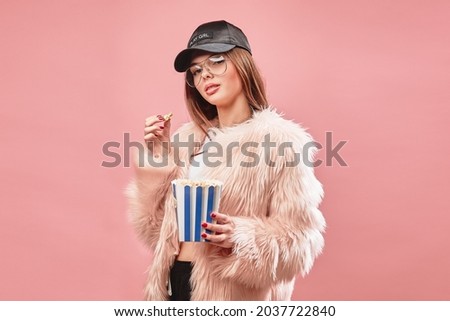Attractive brunette girl in faux pink fur holding popcorn in hand. Wearing in black shorts white top black cap and glasses. Looking at the camera. Studio pink background.