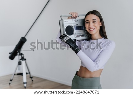 Attractive brunette hispanic girl in sportswear standing at home holds old fashion audio recorder using  cyber arm. Young fit woman with artificial hand happy to be active. Orthopaedics and hi tech.