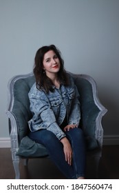 Attractive Brunette In Jeans And Denim Jacket Sitting On Grey Chair. Studio Shoot. 