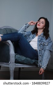 Attractive Brunette In Jeans And Denim Jacket Sitting On Grey Chair. Studio Shoot. 