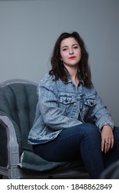 Attractive Brunette In Jeans And Denim Jacket Sitting On Grey Chair. Studio Shoot. 