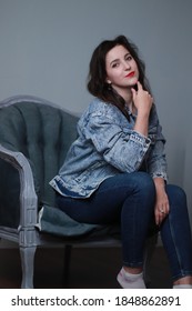 Attractive Brunette In Jeans And Denim Jacket Sitting On Grey Chair. Studio Shoot. 