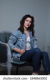 Attractive Brunette In Jeans And Denim Jacket Sitting On Grey Chair. Studio Shoot. 