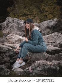 Attractive Brunette Wearing A Jeans Outfit And A Black Cap, Sitting On A Rock In The Nature Looking At Something, Caught In The Moment Of Genuine Pure Beautiful Laughter, Making A Funny Expression