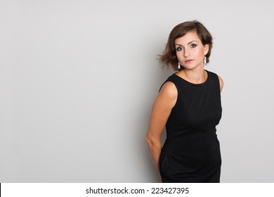 Attractive Brunette Woman In A Black Dress On A Background Of A Gray Wall. Woman 36 Years Old.